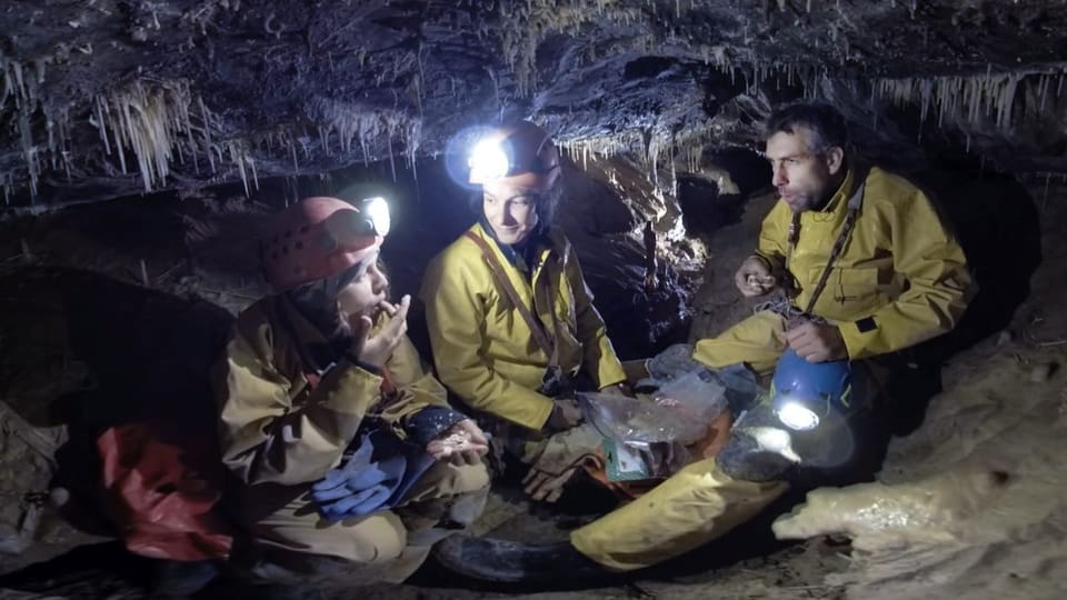 Zwei Frauen und ein Mann mit Helmen und in Arbeitskleidung sitzen in einer Höhle auf dem Boden und essen.