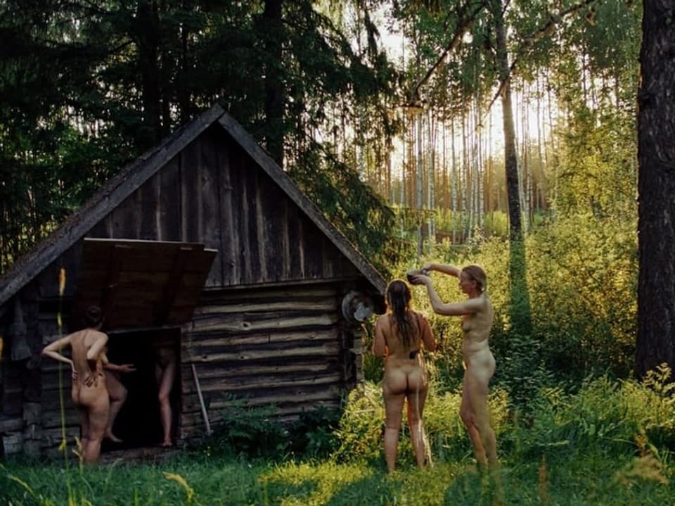 Mitten im Wald steht eine kleine Holzhütte. In, vor und neben der Hütte stehen mehrere nackte Frauen.