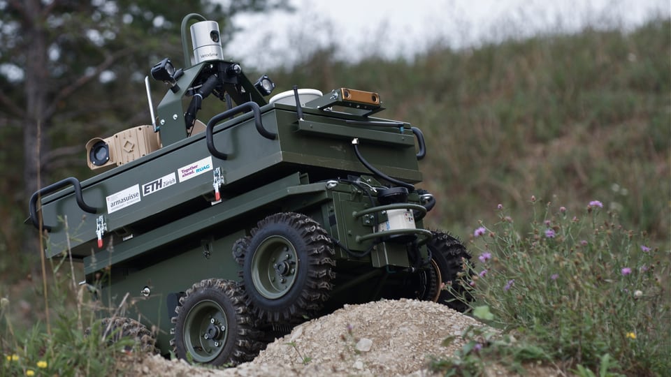 Ein panzerähnliches, dunkelgrünes Fahrzeug mit einem Minikameraaufsatz fährt durch unwegsames Gelände.