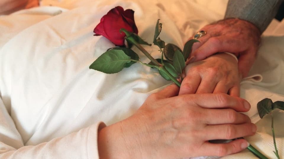 Die gefalteten Hände einer verstorbenen Person umfassen eine Rose.
