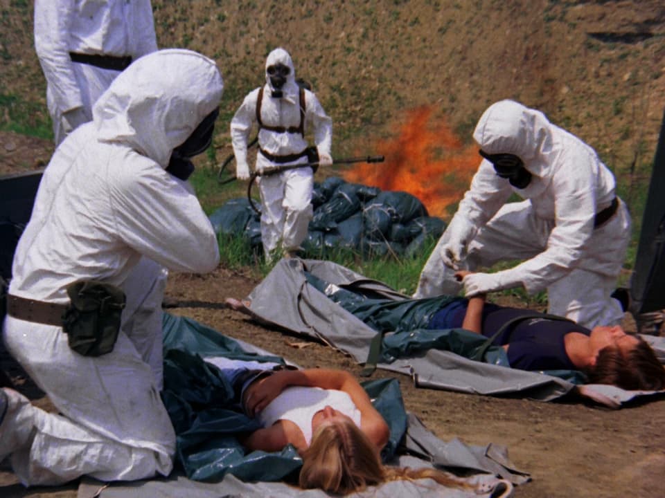 Männer in weisser Schutzkleidung verpacken zwei Menschen in Leichensäcke, im Hintergrund brennt ein Feuer.