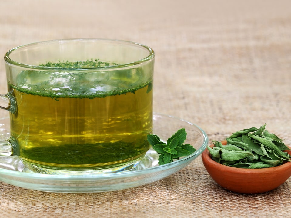 Tasse Tee, darin und daneben getrocknete Stevia-Blätter.