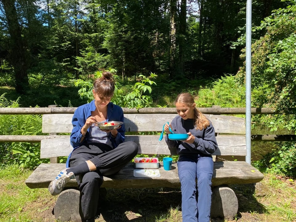 Frau und Mädchen essen in der Sonne auf einer Bank im Wald. 