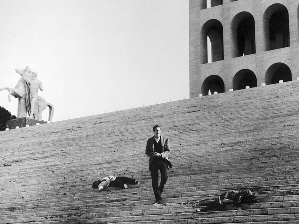 Schwarzweissbild: Ein Mann steht einsam auf einer grossen Trepp, auf der zwei Leichen liegen.