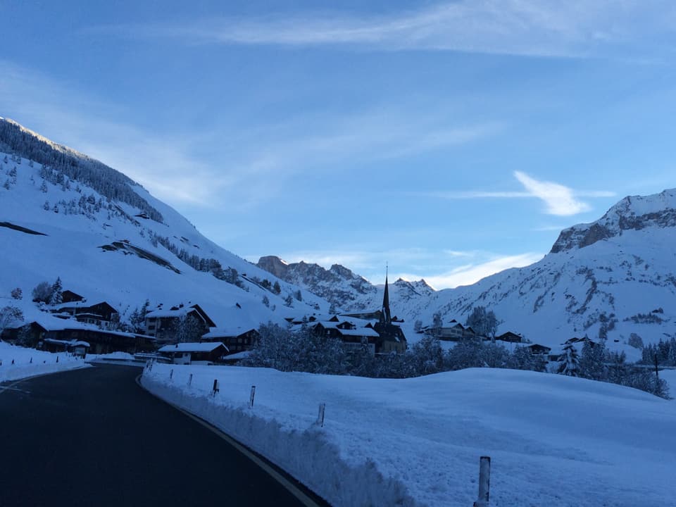 Es ist Dämmerung, der Himmel blau, auch sonst alles in Blautönen. Ein Dorf in einem Alpental, der Blick ist von einer Strasse aus, die ins Dorf führt. Alles ist unter viel Neuschnee begraben. 