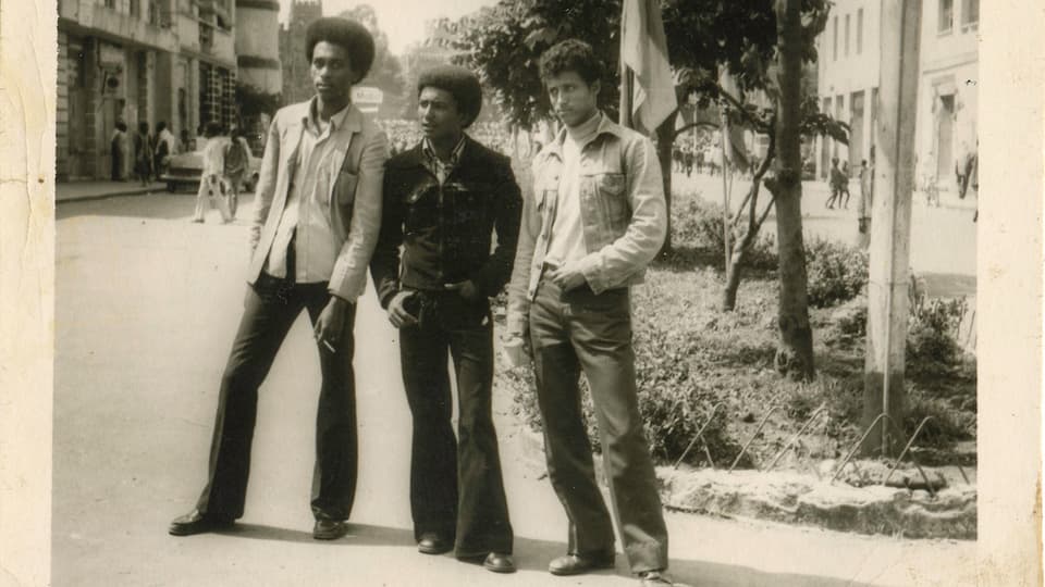 Drei Männer stehen in 70er-Kleidern auf einer Strasse.