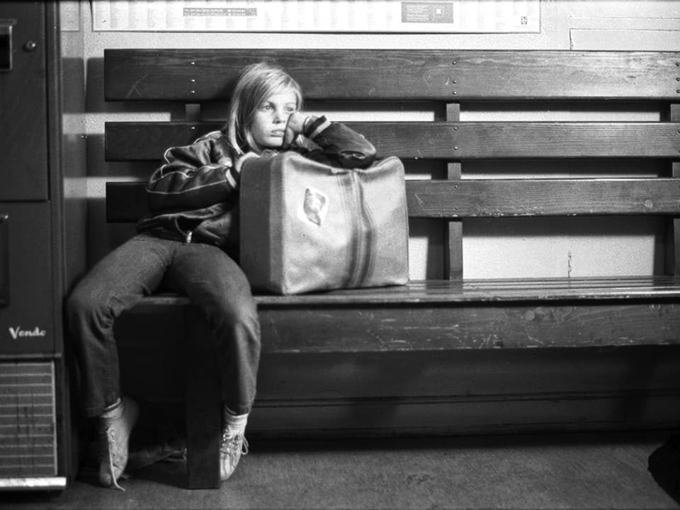 Schwarzweissbild: Ein Mädchen mit gelangweiltem Gesichtsausdruck lehnt sich auf einer Bank gegen einen Koffer.
