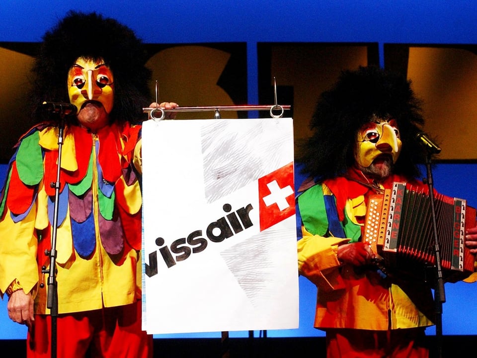 Zwei Fastnachter haben sich bunt kostümiert und halten ein Plakat mit dem Swissair-Schriftzug in die Höhe.