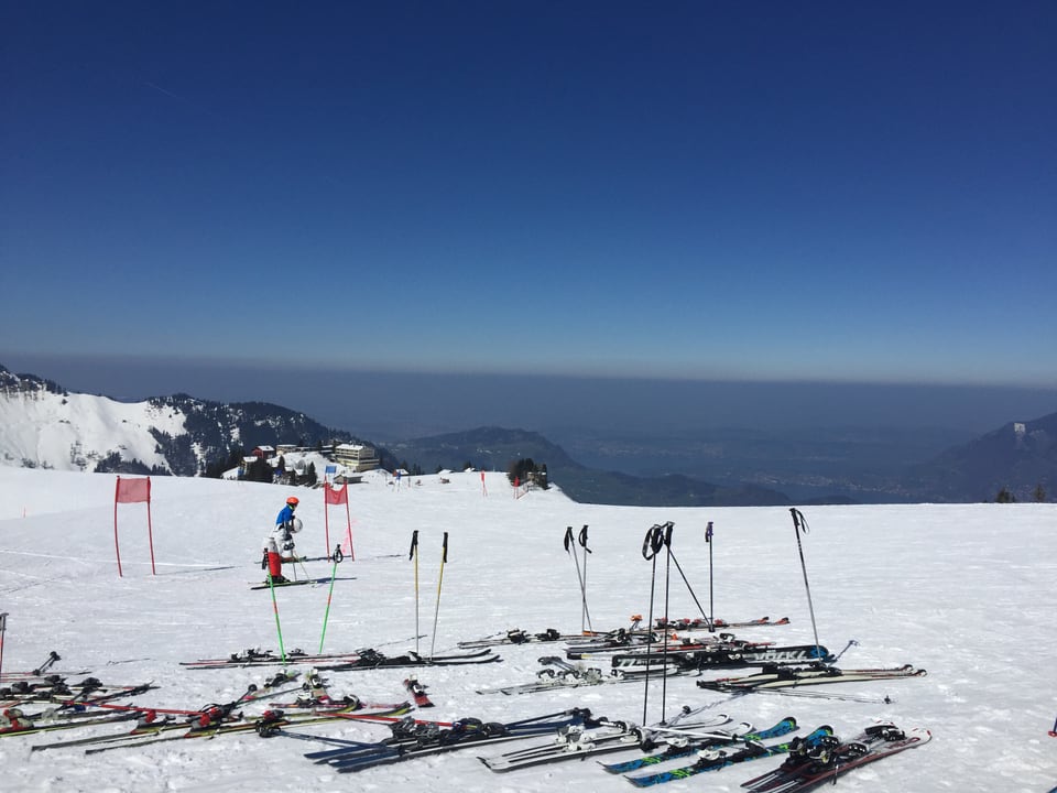 Blick ins Mittelland. Im Vordergrund sind zahlreiche Skier und Skistöcke parkiert. Über dem Mittelland liegt ein dunkler Streifen, Dunst. Darüber ist der Himmel dunkelblau.