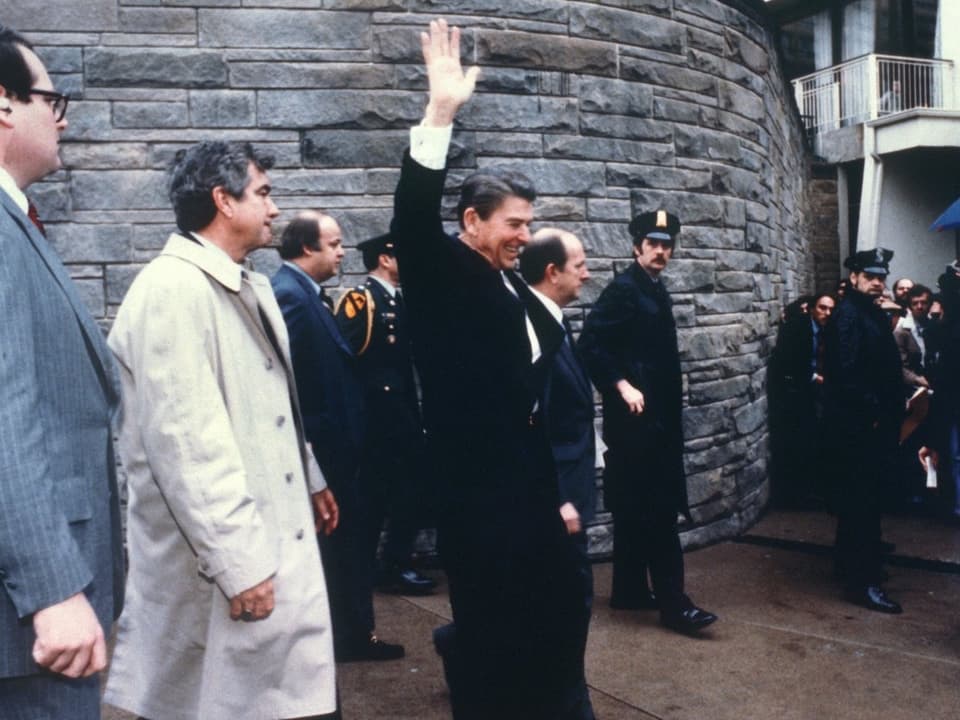 Ronald Reagan winkt kurz vor den Schüssen auf ihn vor dem Hotel Hilton in Washington, rundherum Begleitpersonen, mehrere Polizisten.