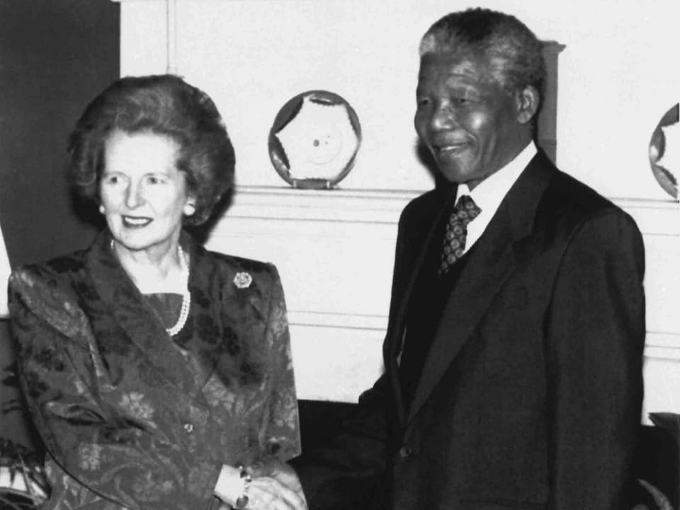 Thatcher und Mandela schütteln sich die Hand und schauen dabei in eine Kamera