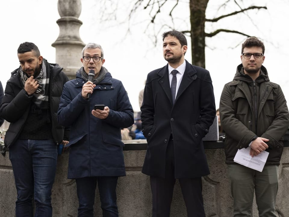 Vier Männer stehen in einer Reihe nebeneinander. Ein Mann hält ein Mikrofon und spricht.