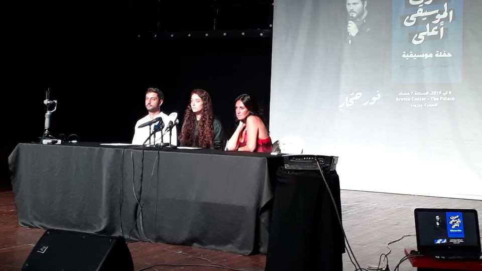 Zwei Frauen und ein Mann sitzen hinter einem Tisch mit Mikrophonen auf einer Bühne.
