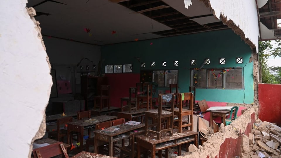 Eine Wand der Schule ist so kaputt, dass man direkt ins Klassenzimmer, mit vielen Holzstühlen und Holztischen, sieht. 