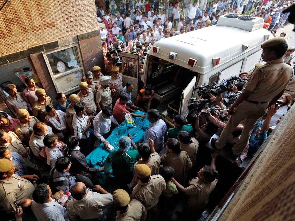 Sicherheitsbeamte schirmen eine Tragbahre ab, die in die Ambulanz geschoben wird. Rundherum drängen Journalisten und weitere Personen.