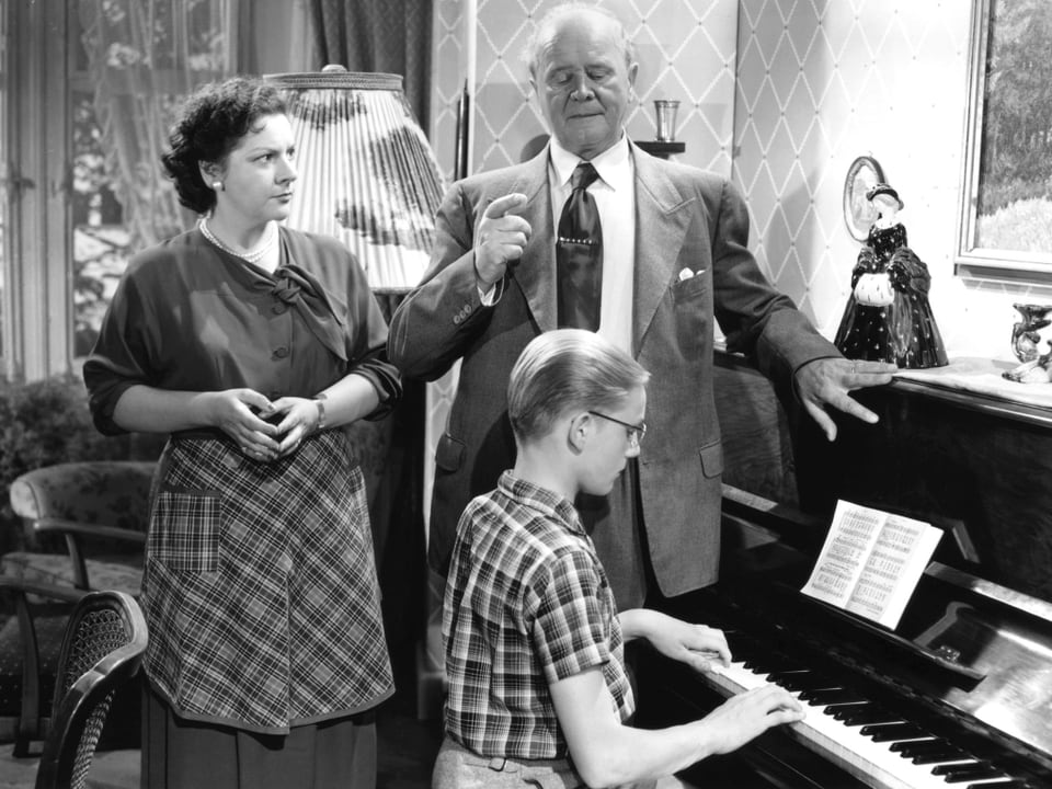 Ein junger Mann mit Brille spielt an einem Klavier. Ein älterer Mann steht neben ihm und zeigt mit dem Zeigefinger den Tackt an. Eine Frau mit Schürze steht neben den beiden und blickt zum Mann.