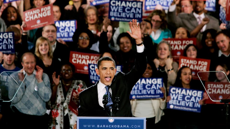Obama spricht am Rednerpult, hinter ihm Menschen mit Plakaten.