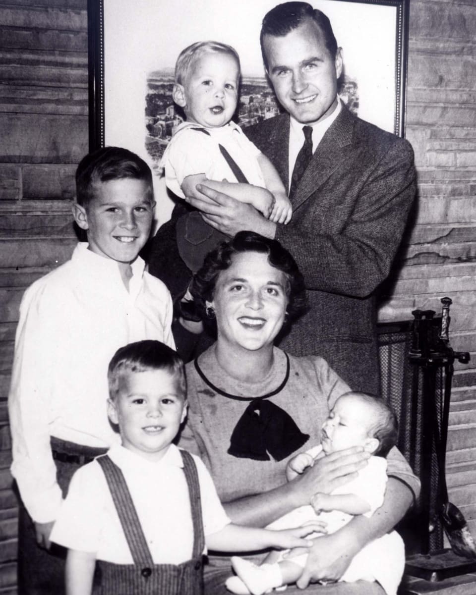 Ein altes Familienporträt. Barbara Bush sitzt auf einem Stuhl und hält ein Baby in ihren Armen. Vater Bush steht hinten und hält Neil im Arm.