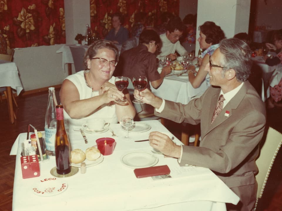 Älteres Ehepaar sitzt an Tisch in Restaurant der 1970er-Jahre und prostet sich mit einem Glas Wein zu.