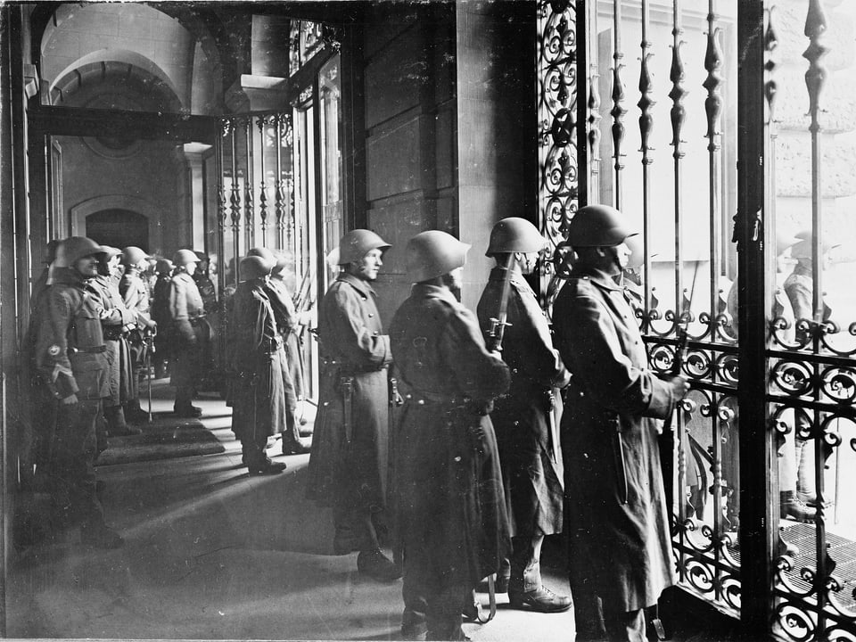 Hinter geschlossenen Gittern stehen Soldaten und blicken auf den Bundshausplatz.