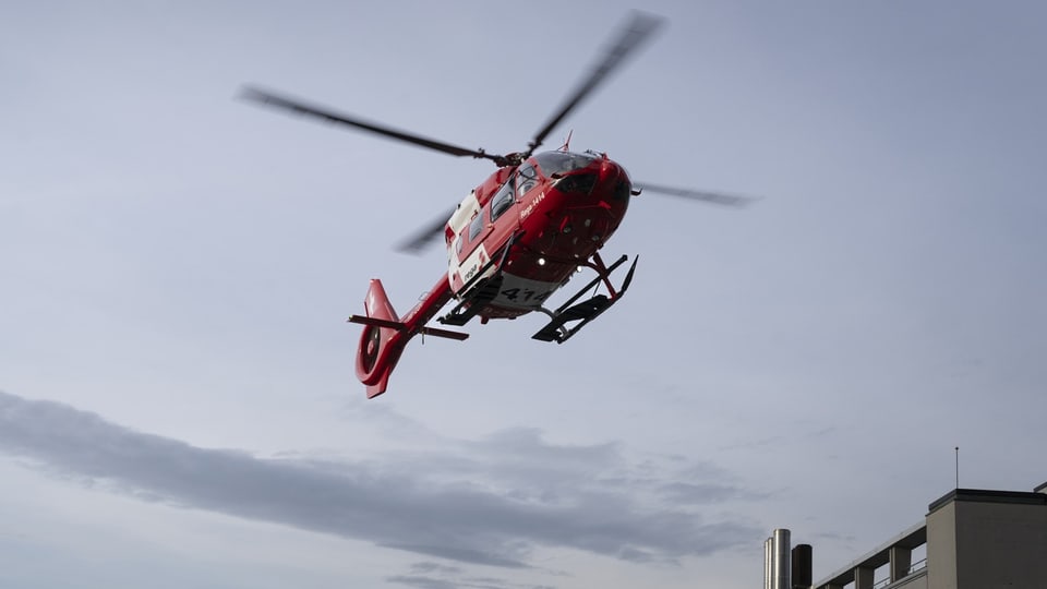 Rega-Helikopter in rot weiss, in der Luft, dahinter braunes Hochhaus.