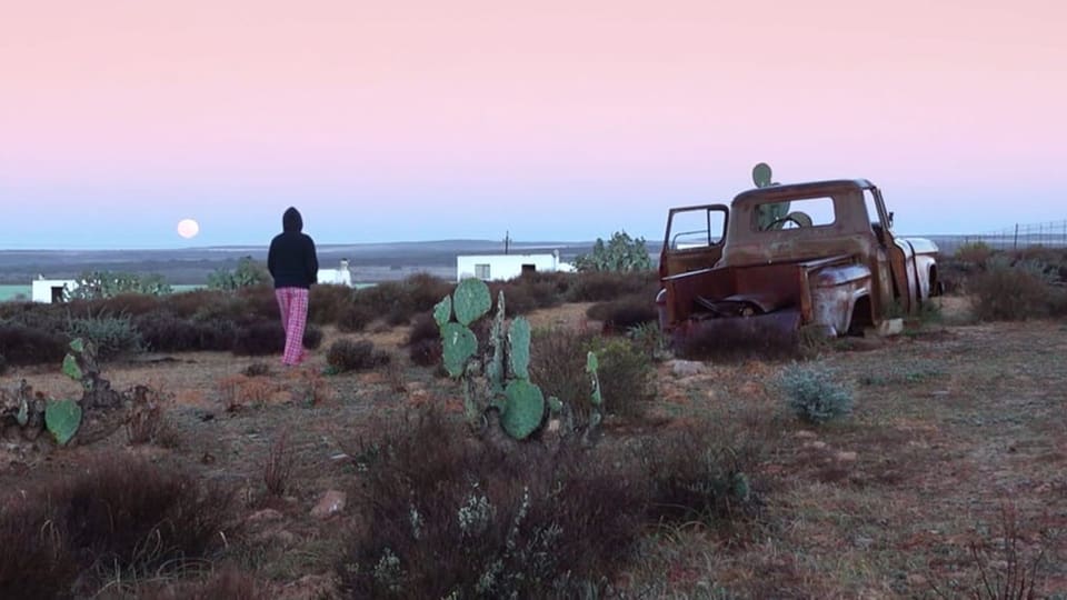 Eine Frau mit Kapuzenpullover läuft in der Wüste neben einem Schrottauto.