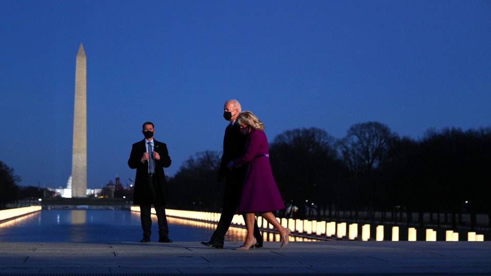 Mann und Frau schreiten über Platz. Leuchtstäbe am Boden erinnern an die Verstorbenen. Im Hintergrund der Obelisk von Washington.