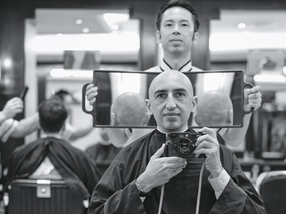 Ein Mann sitzt mit Glatze in einem Coiffeurladen, hinter ihm der Friseur mit einem grossen Spiegel in der Hand.