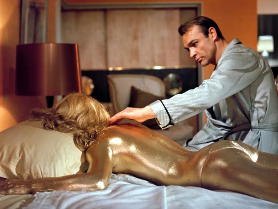 Ein Mann im Morgenmantel sitzt auf dem Bett und streicht mit der Hand über eine weibliche Figur aus Gold, die auf dem Bauch liegt.