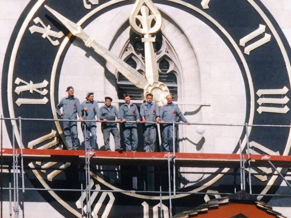 Sechs Männer stehen auf einem Gerüst vor der Uhr des St.Peters.