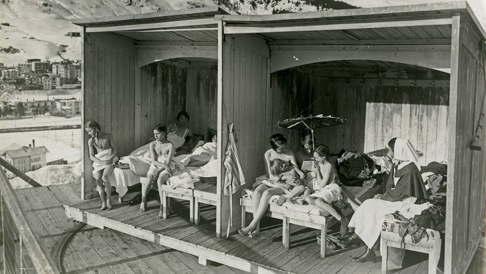 eine schwarz-weiss Fotografie von Holzblöcken im Freien und Frauen, die sich darin sonnen