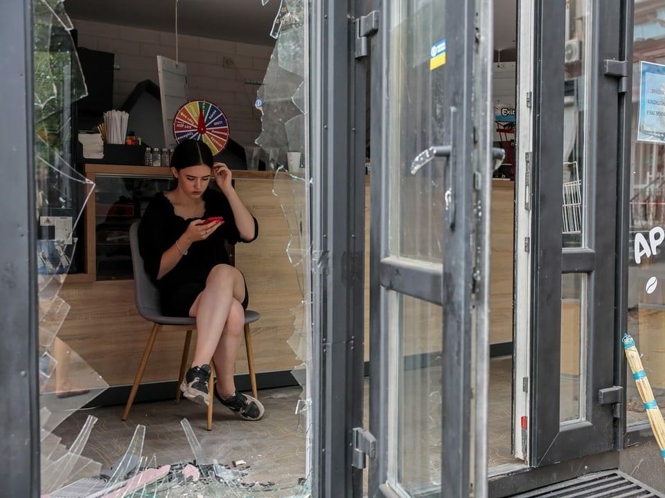 Eine Frau sitzt auf einem Stuhl und schaut auf hier Handy. Vor ihr ist eine zerbrochene Glastüre. 