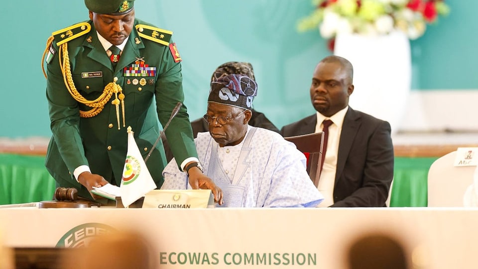 älterer Mann mit Kopfbedeckung an Tisch, ein Mann in grünem Militäranzug beugt sich über ihn.
