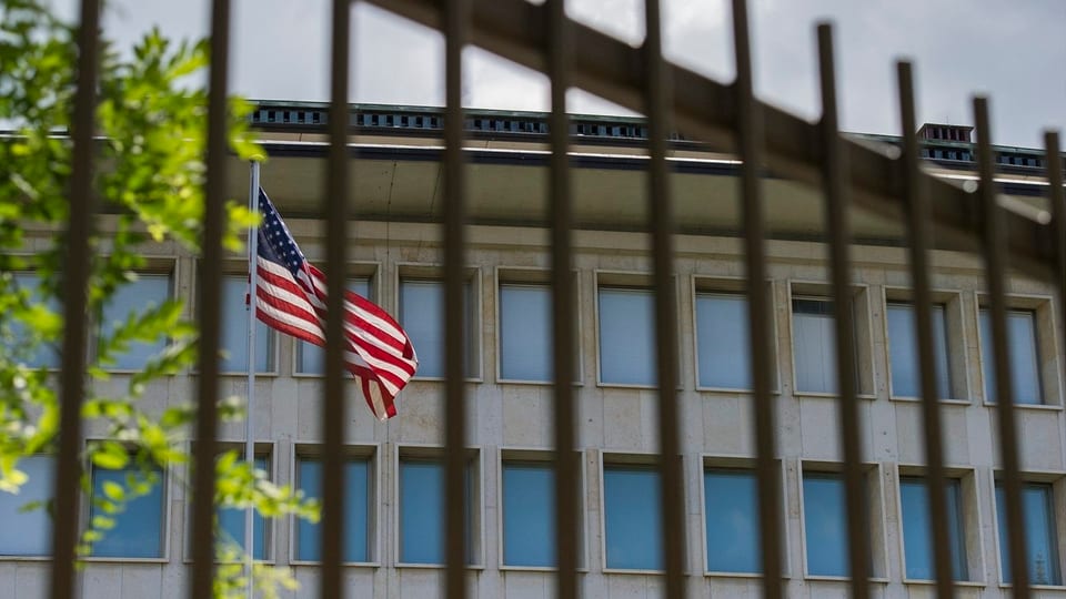 Gebäude hinter einem Eisenzaun, davor eine US-Flagge auf einem Fahnenmast.