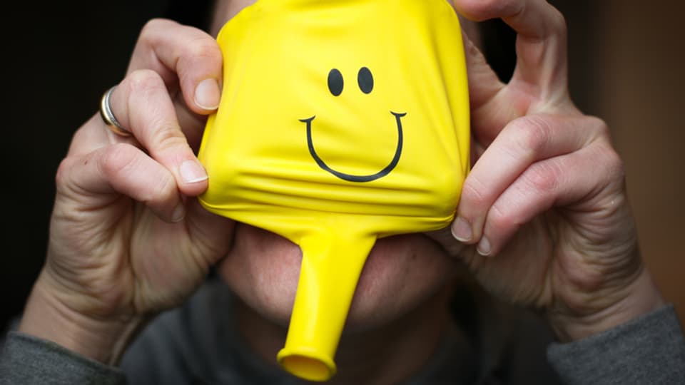 Eine Frau hält einen Ballon vor das Gesicht. Es ist ein gelber Ballon mit einem Smilie-Gesicht.