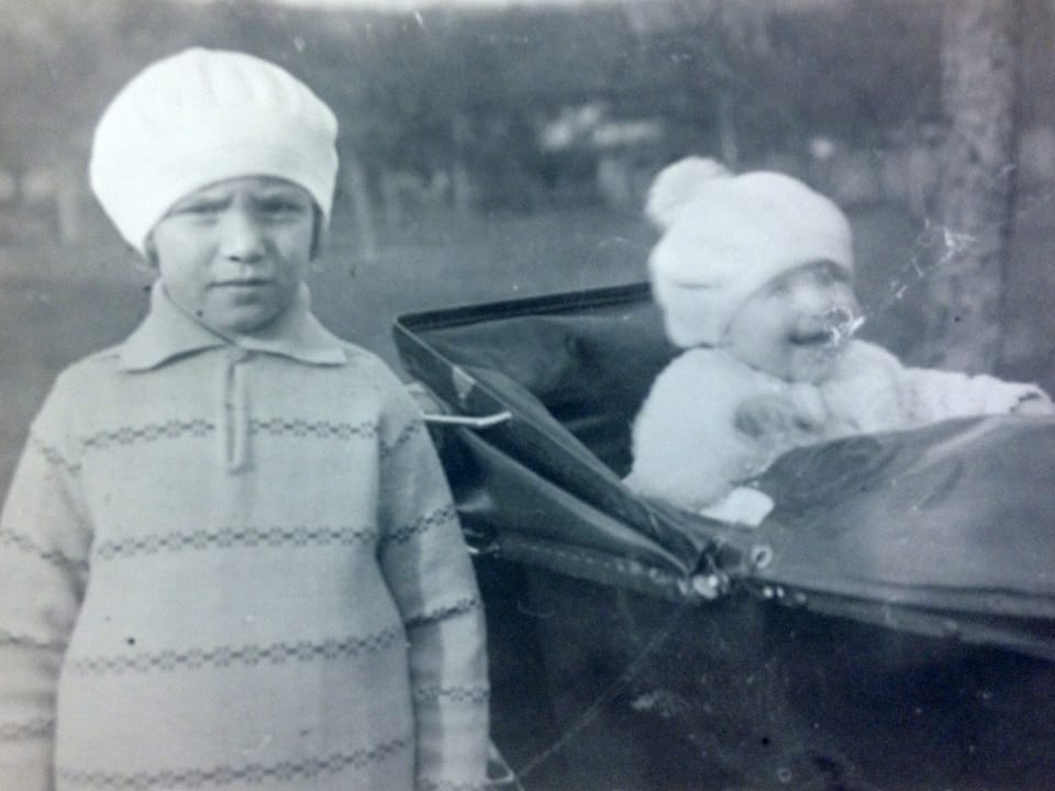 Ein kleines Mädchen mit Mütze steht neben einem Kinderwagen, indem ein Kleinkind sitzt.
