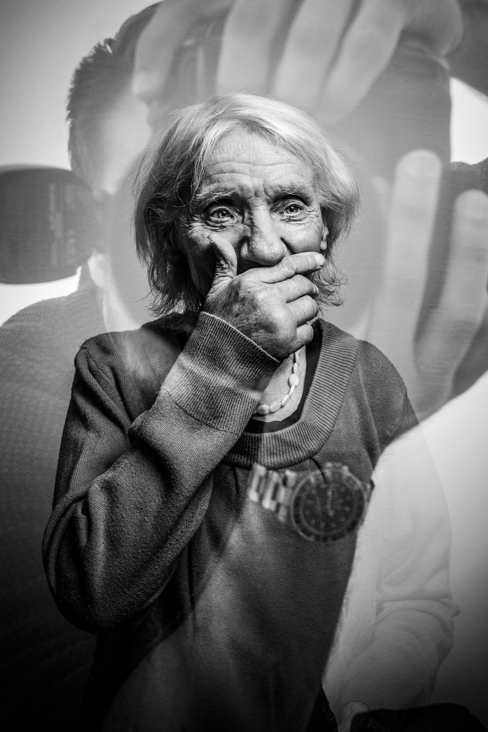 Portrait einer älteren Dame, die sich eine Hand vor den Mund hält. Im Bild spiegelt sich der Fotograf.