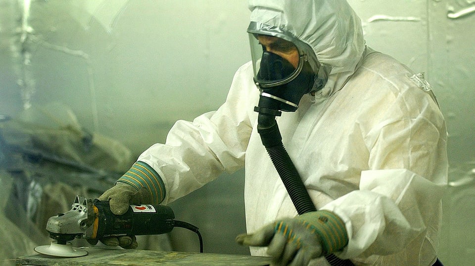 Ein in Schutzkleidung gehüllter Arbeiter schleift ein asbestverseuchtes Werkstück.