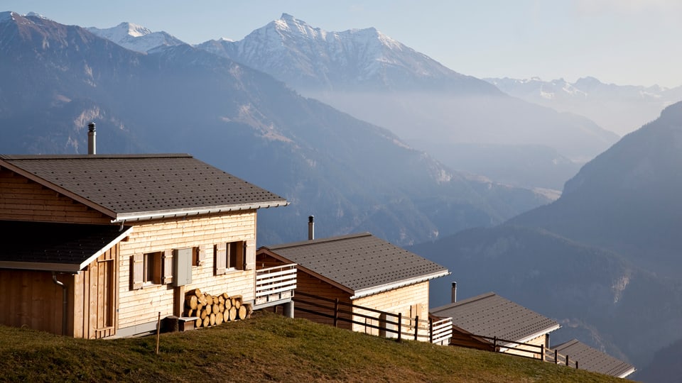 Drei Holzhäuser auf einer Bergkuppe. Im Hintergrund Berge.