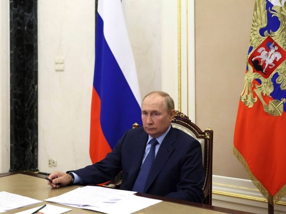 Ältere Mann in Anuzug (Putin) sitzt an Schreibtisch vor russischer Flagge