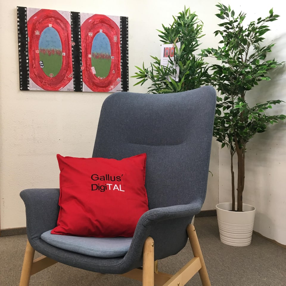 Ein grauer Sessel mit rotem Kissen steht in einer Ecke, an der Wand hängt abstrakte Kunst, hinter dem Sessel hat es zwei meterhohe grüne Pflanzen