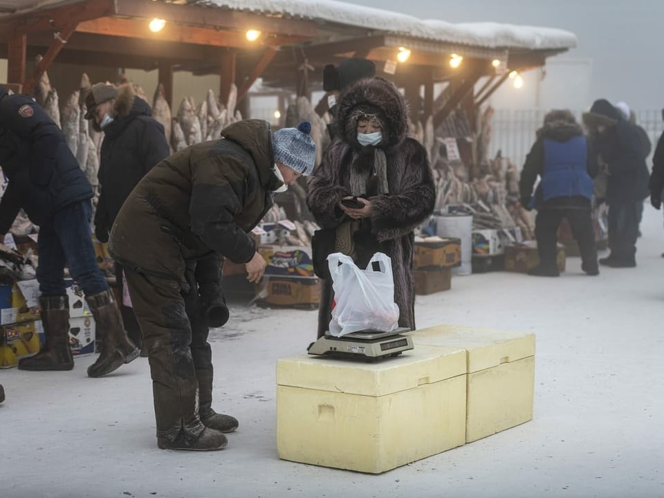 Ein Mann wiegt Ware auf einer Waage, im Hintergrund gefrorene Fische auf einem Markt in Jakutsk, alle tragen Pelz.