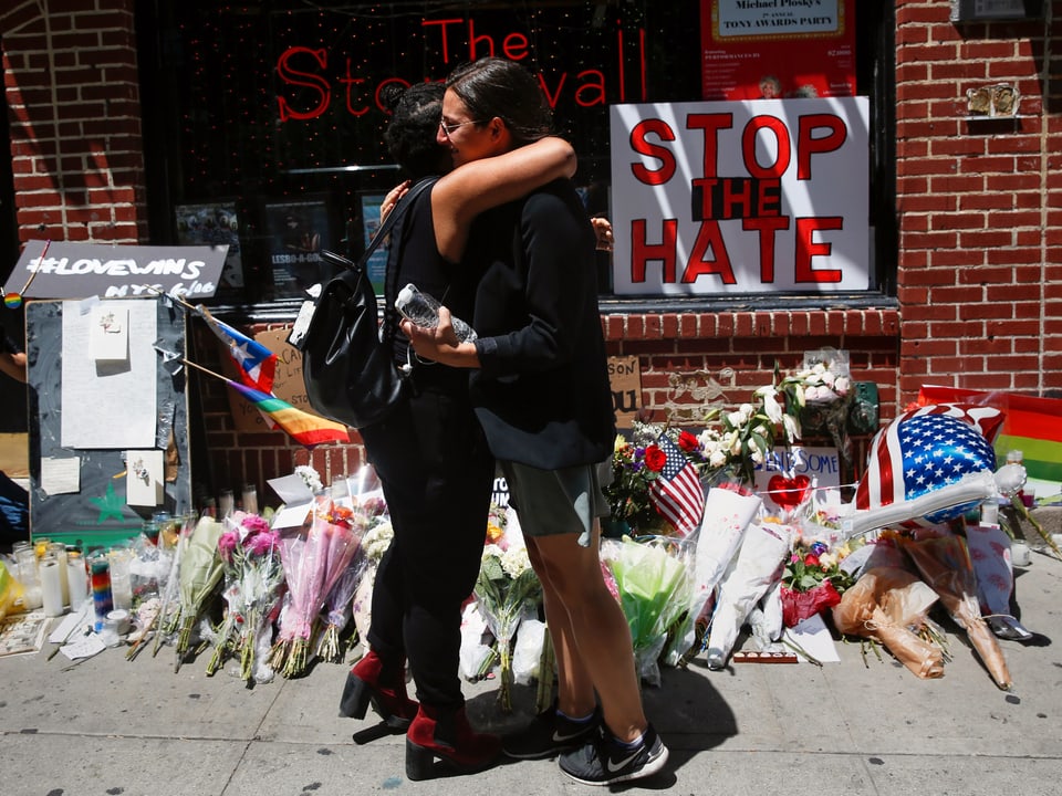 Zwei Frauen umarmen sich vor der Bar "The Stonewall Inn". Am Boden vor der Bar liegen viele Rosensträusse. An der Wand ein Schild "Stop the Hate".