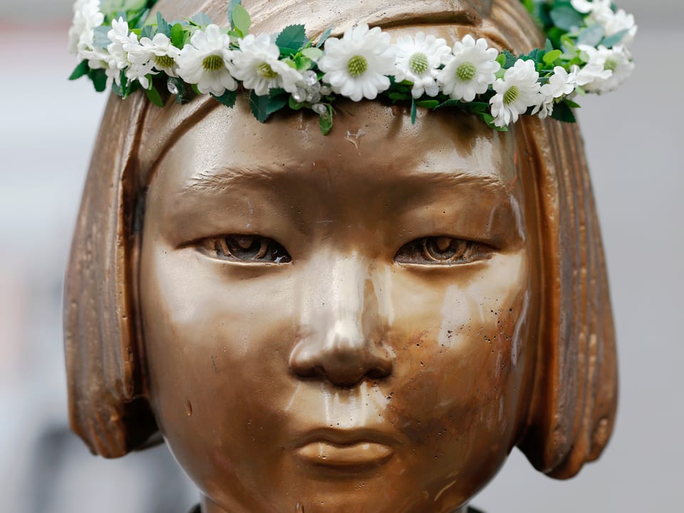 Kopf einer Bronzestatue eines asiatischen Mädchens, jemand hat einen Kranz aus weissen Büten auf den Kopf gelegt.
