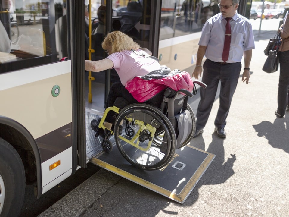 Eine Frau auf einem Rollstuhl fährt auf einer Rampe in einen Bus. Daneben steht der Chauffeur.