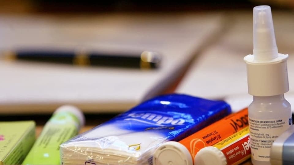 Auf einem Tisch liegen verschiedene Medikamente gegen Erkältung, ein Taschentuchpäckchen sowie ein Block und Stift.