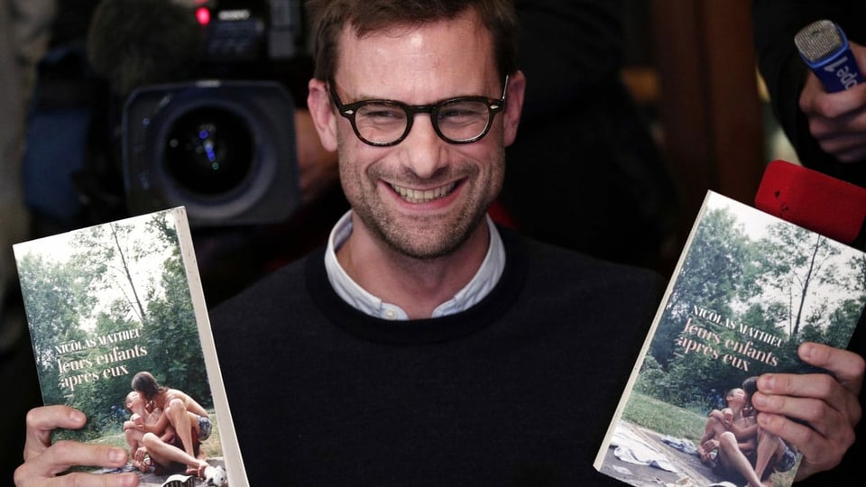 Ein Mann mit markanter Brille hält strahlend zwei Bücher in die Kamera.