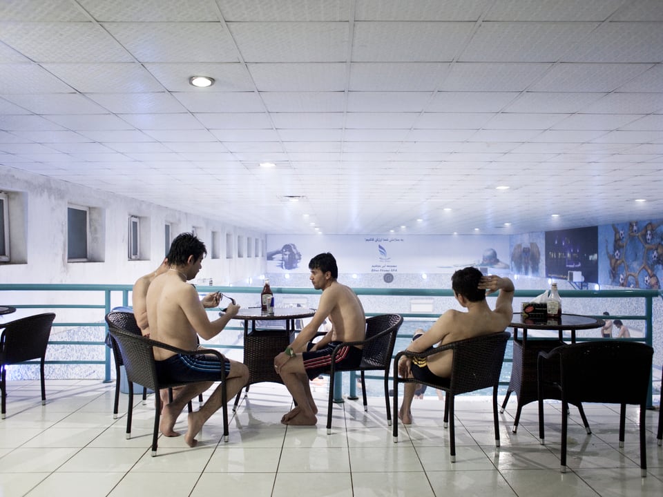 Männer sitzen mit nacktem Oberkörper auf schwarzen Stühlen in einem Hallenbad.
