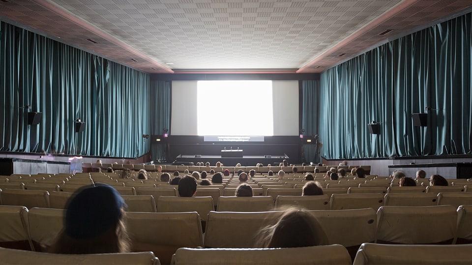 Blick von hinten in den Kinosaal, auf der Leinwand läuft ein Film, in den Stühlen sitzen vereinzelt Zuschauer.