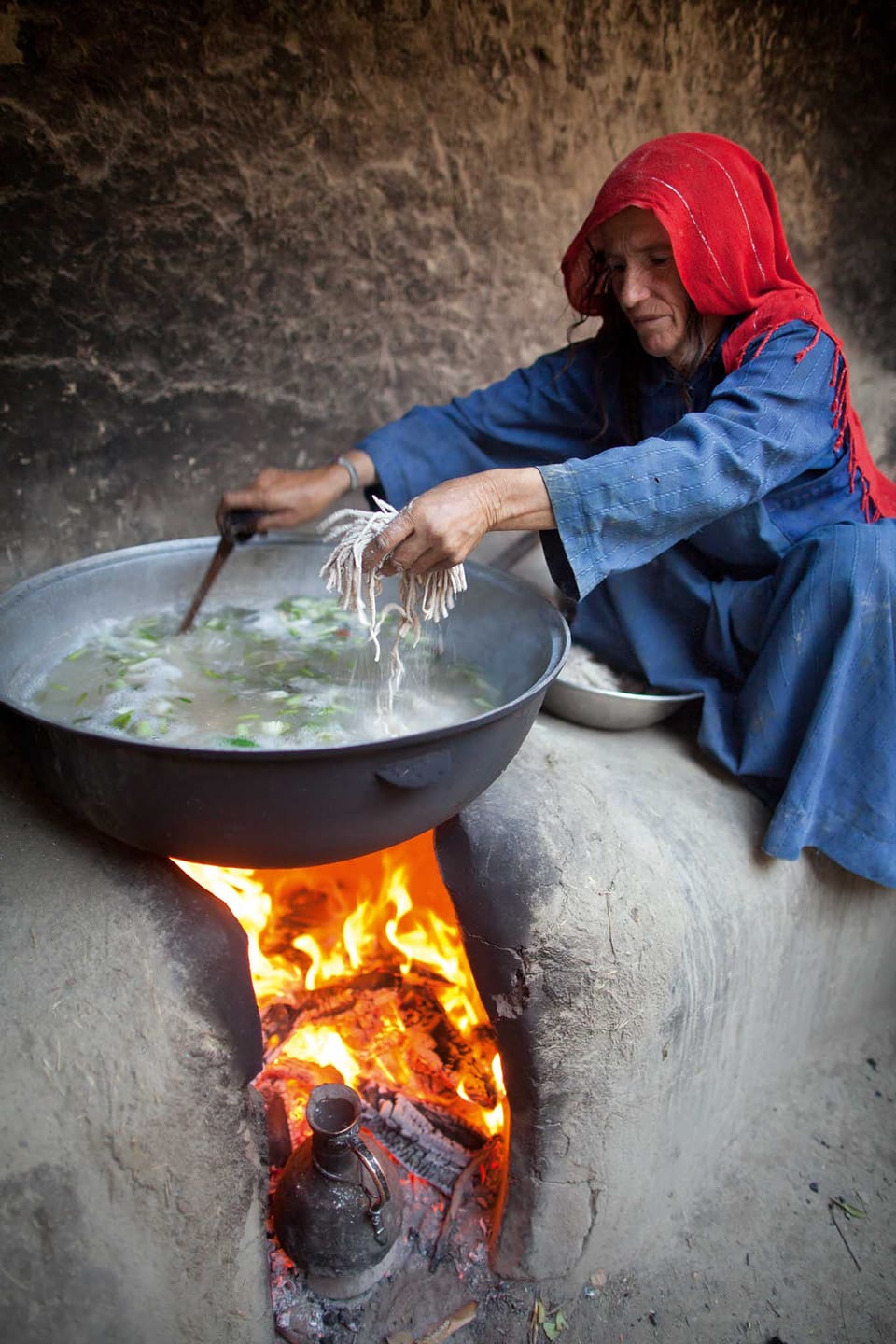 Eine Frau gibt Nudeln in einen grossen Suppentopf, der auf einem Steinofen über einem Feuer steht.